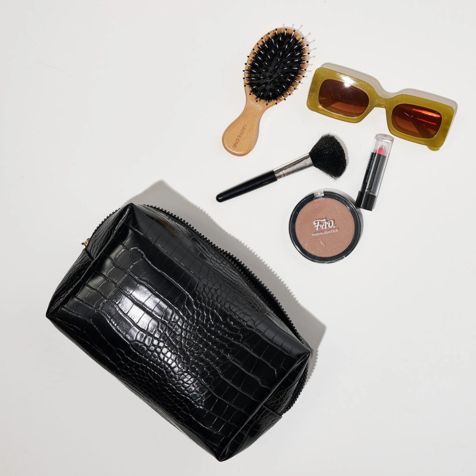 Black crocodile-patterned clutch with makeup items including a brush, compact, and sunglasses on a light gray background.