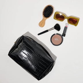 Black crocodile-patterned clutch with makeup items including a brush, compact, and sunglasses on a light gray background.