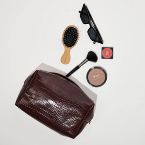 Brown crocodile-patterned clutch with makeup items including a brush, compact, and sunglasses on a light gray background.